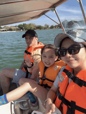 A mother and father sit on either side of their daughter in a boat. All three are wearing orange life jackets and smiling at the camera.