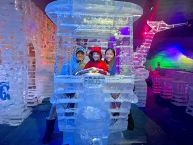 A father, mother, and their young daughter pose for a photo inside an ice tuk-tuk. They're all wearing warm jackets, and other ice sculptures are visible around them.