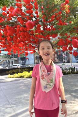 A young girl wearing a pink shirt and a scarf around her neck smiles happily while standing on a shady sidewalk under a large tree decorated in red for Chinese New Year.