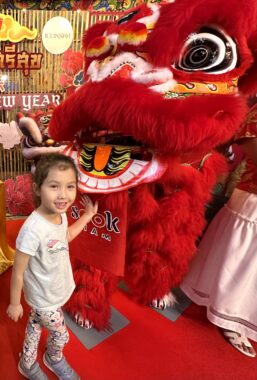 A young girl smiles and pats a bright red Chinese New Year lion with her left hand during a traditional dance. 