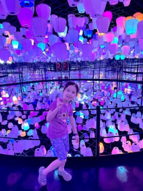 A 7-year-old girl smiles for a photo inside some sort of art installation. The room is dark and filled with colorful lanterns.
