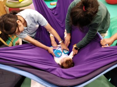 A baby girl lies on her back in a large sling suspended a few feet above the ground. Two adults are working with her on some sort of early intervention therapy.