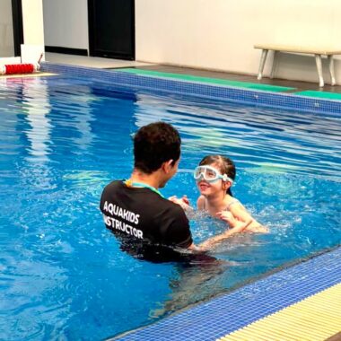 A young child wearing goggles is led in a pool by a swimming instructor.