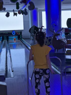 A young child watches as a bowling ball rolls toward the pins.