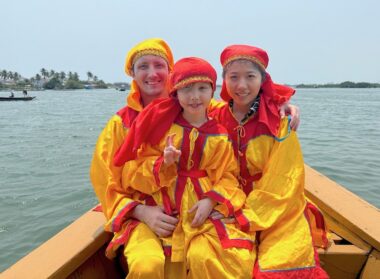 Two adults and a child are shown in bright yellow and red outfits sitting in a boat in a river.