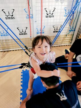 A child is supported by ropes as she undergoes a session of physical therapy on a mat.
