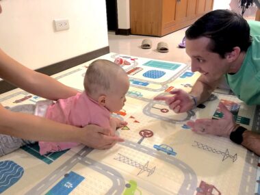 A baby lies on a rug on a floor opposite a man who gestures toward her.