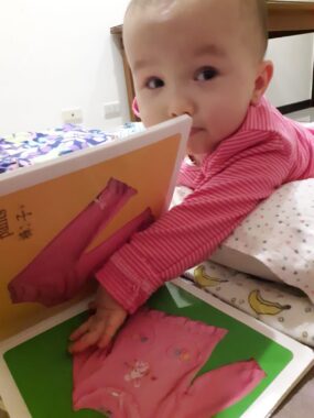 A baby in bed, lying on her stomach, extends her left arm and hand into a children's book that has pink clothing matching the onesie that she's wearing.