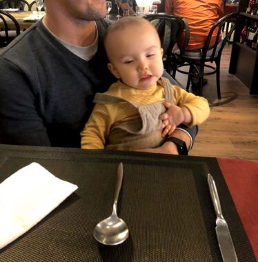 A close-up of a baby sitting on her father's lap at a restaurant table. The father's face isn't in the frame, but the focus of the photo is the baby staring at a large spoon on the table. 