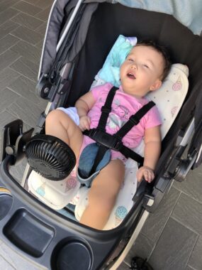 A toddler is strapped in a baby carrier that has a small, black fan attached to the front of it. 