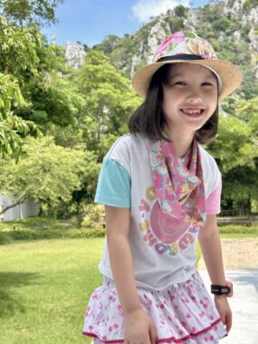 A 7-year-old girl smiles while standing outdoors on a sunny day, among trees and mountains. She's wearing a colorful outfit and fedora.