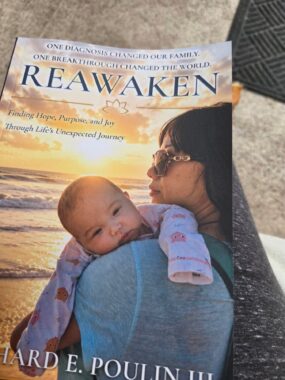 The cover of a book titled "Reawaken" is pictured, featuring a photo of a woman holding a baby on a beach at sunset.