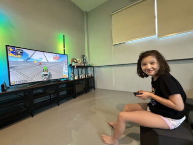 An 8-year-old girl sits on the edge of a couch holding a video game controller. The TV in front of her shows her in the middle of a Mario Kart race, though she's turned to the side and smiling at her dad, who's taking the photo.