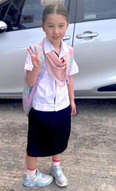 A young girl dressed in a school uniform waves as she stands in front of a parked car.