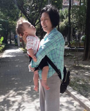 An older woman standing outside in a park holds an infant in her arms, with her right hand cupping the back of the child's head