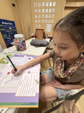 A young girl sits at a table and writes in some sort of school workbook. 