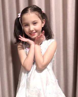 A young girl dressed in a white dress smiles and cups her face in her hands.