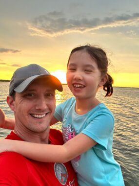 A child wraps her arms around a man's shoulders as they smile for a photo in front of an expanse of water at sunset.