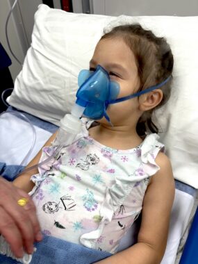 A young girl reclines in a bed as she wears a nebulised medicine face mask